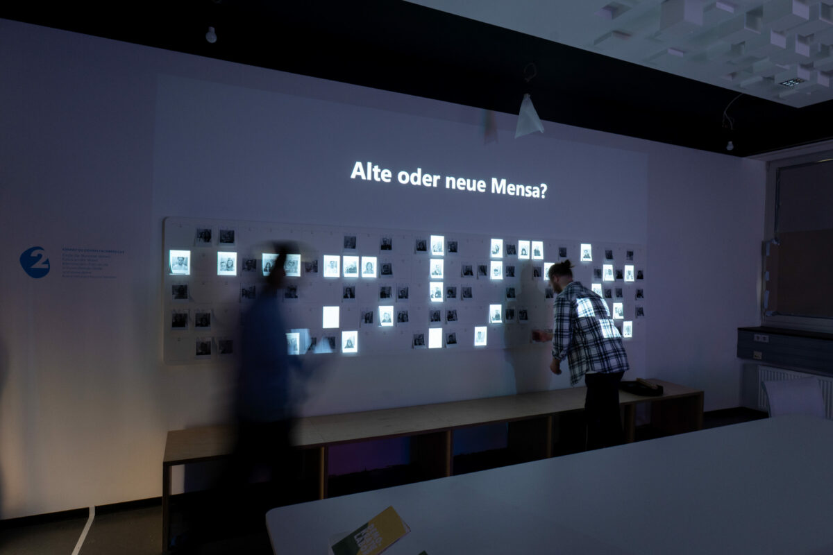Eine Wand mit vielen ausgedruckten Poirtraitfotos und einer überlagernden Projektion. Über den Bildern wird gerade die Frage "alte oder neue Mensa" projiziert und einzelne Personen sind dazu angeleuchtet