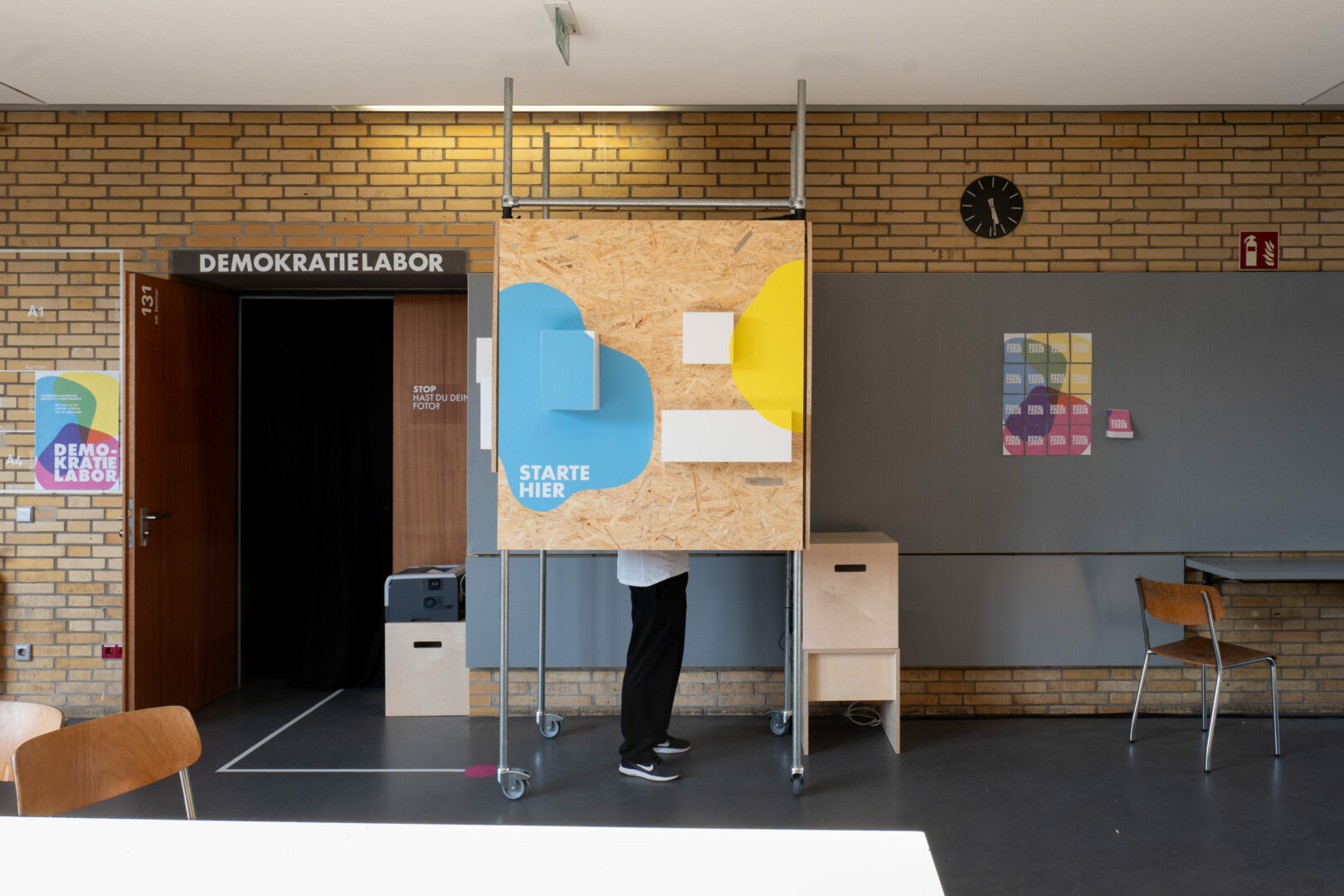 Eine Person steht hinter einer mobilen Holzinstallation auf Rollen im Eingangsbereich des Demokratielabors. Auf der Vorderseite sind bunte Formen in Blau und Gelb mit dem Schriftzug „STARTE HIER“ angebracht. Links führt eine offene Tür mit schwarzem Vorhang in einen Raum mit der Beschriftung „DEMOKRATIELABOR“. An der Wand hängen farbige Plakate und ein schwarzer Wanduhr zeigt die Zeit. Der Raum wirkt wie ein experimenteller Lern- oder Ausstellungsort.