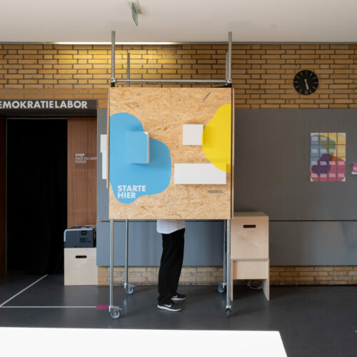 Eine Person steht hinter einer mobilen Holzinstallation auf Rollen im Eingangsbereich des Demokratielabors. Auf der Vorderseite sind bunte Formen in Blau und Gelb mit dem Schriftzug „STARTE HIER“ angebracht. Links führt eine offene Tür mit schwarzem Vorhang in einen Raum mit der Beschriftung „DEMOKRATIELABOR“. An der Wand hängen farbige Plakate und ein schwarzer Wanduhr zeigt die Zeit. Der Raum wirkt wie ein experimenteller Lern- oder Ausstellungsort.