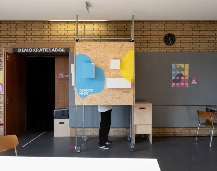 Eine Person steht hinter einer mobilen Holzinstallation auf Rollen im Eingangsbereich des Demokratielabors. Auf der Vorderseite sind bunte Formen in Blau und Gelb mit dem Schriftzug „STARTE HIER“ angebracht. Links führt eine offene Tür mit schwarzem Vorhang in einen Raum mit der Beschriftung „DEMOKRATIELABOR“. An der Wand hängen farbige Plakate und ein schwarzer Wanduhr zeigt die Zeit. Der Raum wirkt wie ein experimenteller Lern- oder Ausstellungsort.